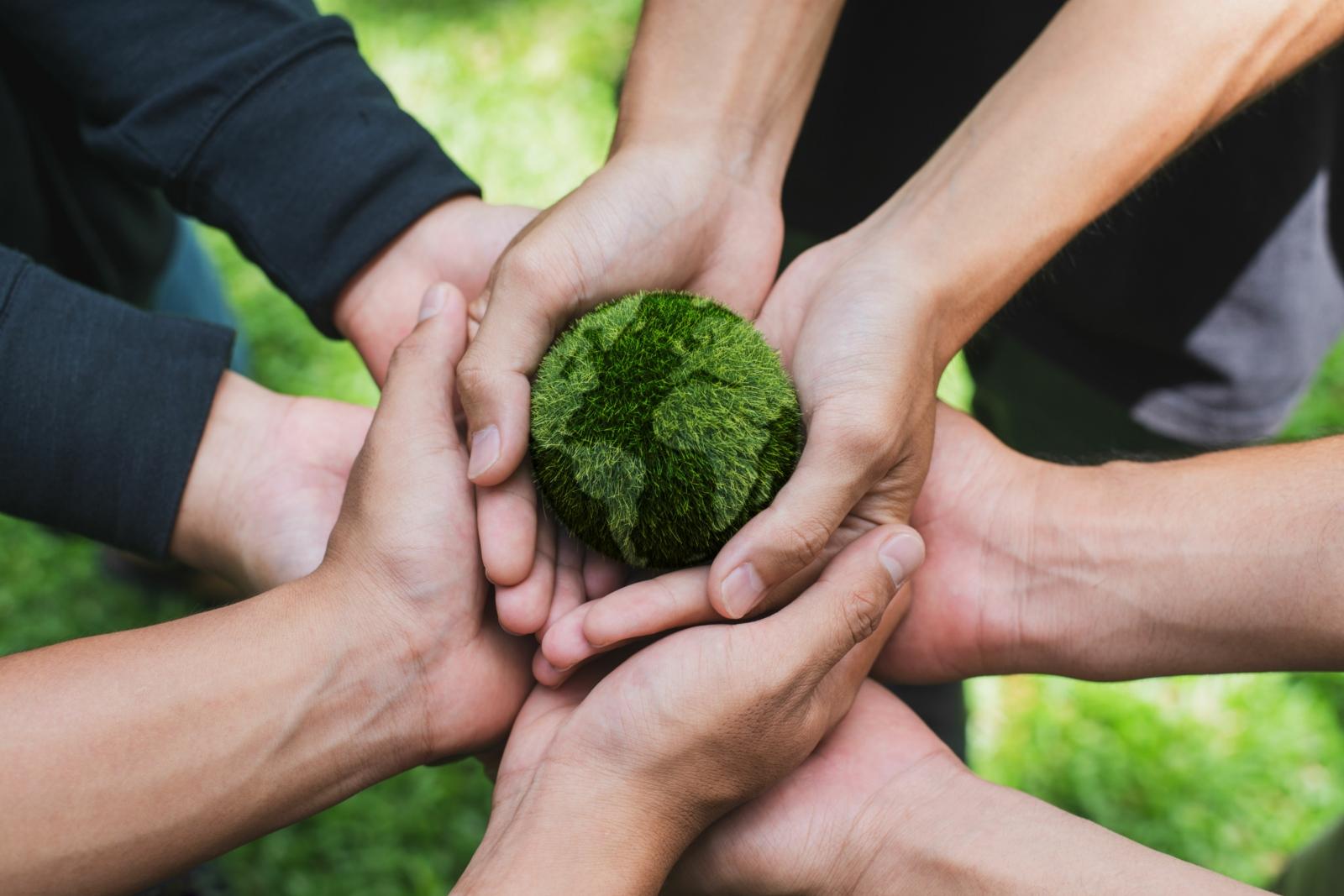 Hände tragen gemeinsame eine Weltkugel. Die Erde ist aus Gras dargestellt.